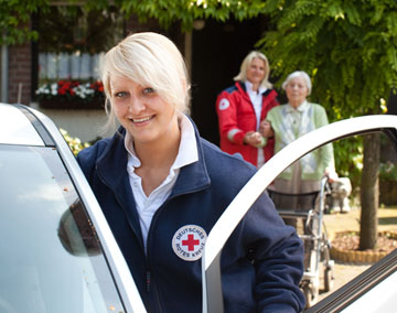 Foto: Eine blonde Pflegekraft steht lchelnd an einem Einsatzwagen.